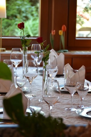 Ein elegant gedeckter Tisch mit Weingläsern, weißen Servietten und roten Rosen in einem Restaurant. Im Hintergrund sind Fenster und Tulpen zu sehen., © Badhotel Restaurant Stauferland
