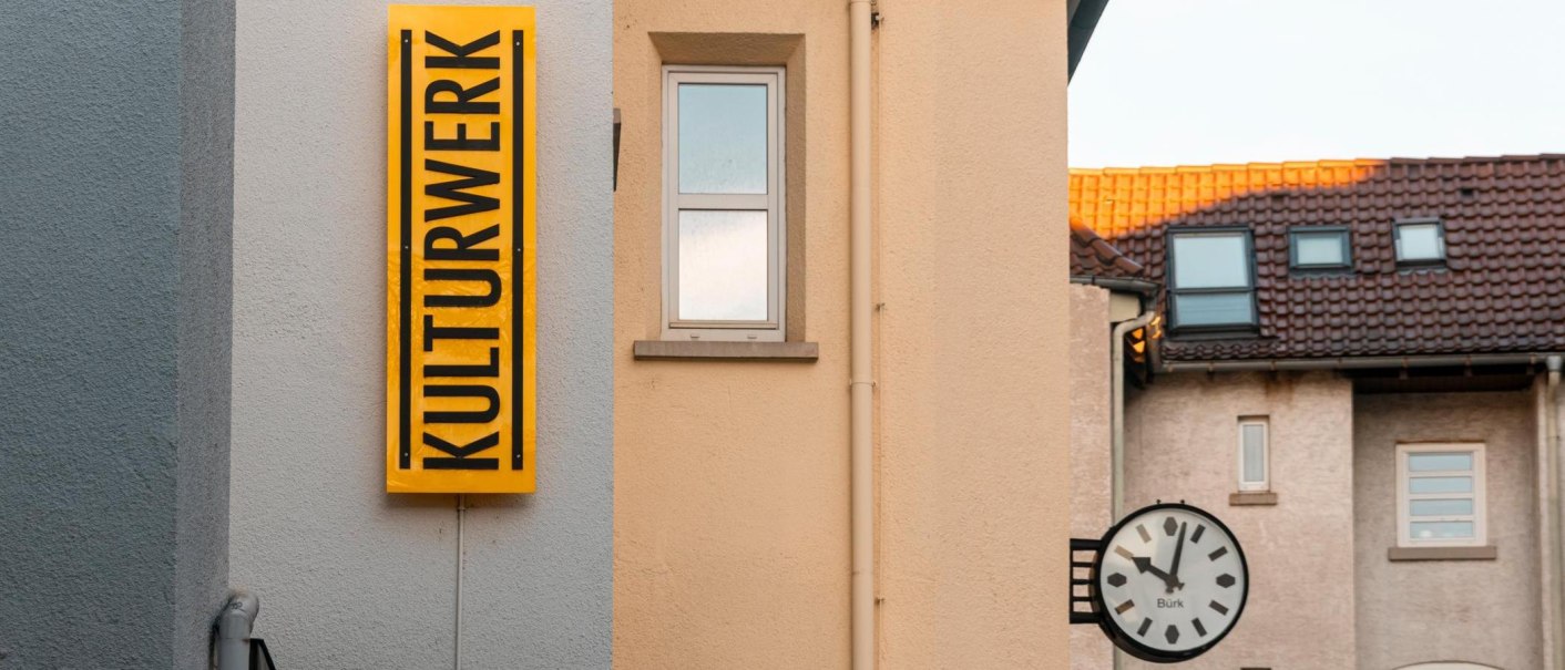 A yellow sign with the inscription 'KULTURWERK' hangs on the wall of a house. Next to it is a window and a clock., &copy; SMG Stuttgart Marketing GmbH - Sarah Schmid