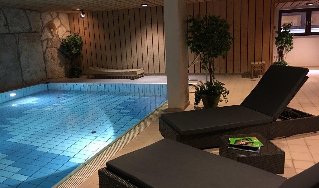 Ein beheizter Indoor-Pool mit blauen Fliesen, umgeben von Liegestühlen und Pflanzen. Die Wände sind aus Holz und Stein., © TOMAS