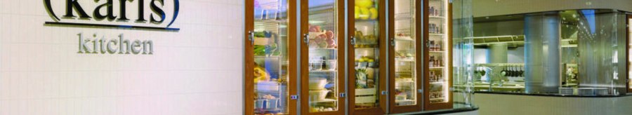 Entrance to "karls kitchen" with illuminated glass cabinets full of food and a view of the modern kitchen., © karls kitchen, Stuttgart