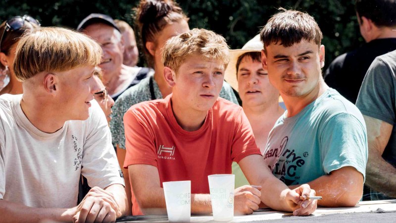 Drei junge Männer in einer Menschenmenge, zwei mit Plastikbechern, einer raucht. Sie wirken konzentriert und sind im Freien., © Pandora Film