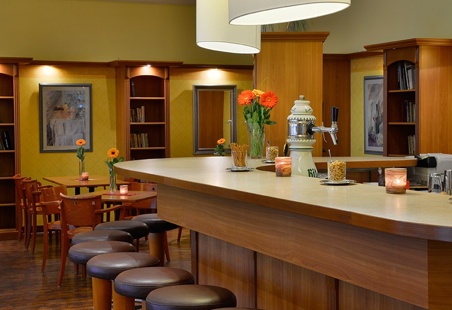 An elegant bar with a wooden counter, bar stools and stylish lighting. Flowers and candles decorate the counter, with tables and shelves in the background., © Amber Hotel Leonberg / Stuttgart An elegant bar with a wooden counter, bar stools and stylish lighting. Flowers and candles decorate the counter, with tables and shelves in the background., © Amber Hotel Leonberg / Stuttgart