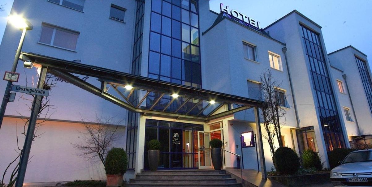 Modernes Hotel mit Glasfassade und beleuchtetem Eingang bei Dämmerung. Ein Straßenschild zeigt 'Lessingstraße'. Ein Auto parkt vor dem Gebäude., © Hotel am Park Modernes Hotel mit Glasfassade und beleuchtetem Eingang bei Dämmerung. Ein Straßenschild zeigt 'Lessingstraße'. Ein Auto parkt vor dem Gebäude., © Hotel am Park