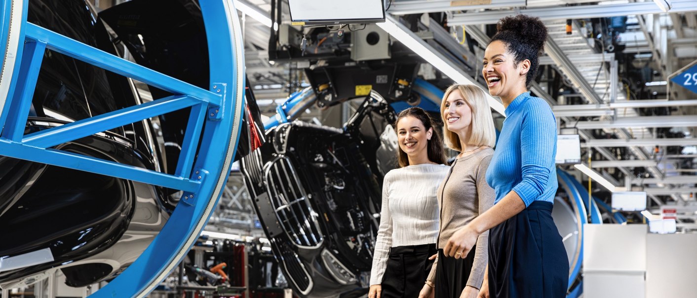 Three women stand smiling in a modern factory hall. Car parts and industrial equipment can be seen in the background., © Mercedes-Benz AG