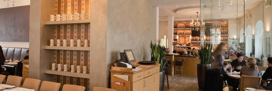 Stylish restaurant with wooden furniture, shelves full of cups and a cozy atmosphere. Guests sit at tables, with a bar in the background., &copy; Aspectus Photographie Stuttgart