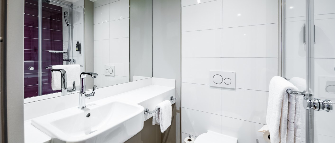 Modernes Badezimmer mit weißem Waschbecken, Toilette und verglaster Dusche. Handtücher hängen an Haltern, helle Fliesen dominieren den Raum., © Premier Inn Modernes Badezimmer mit weißem Waschbecken, Toilette und verglaster Dusche. Handtücher hängen an Haltern, helle Fliesen dominieren den Raum., © Premier Inn