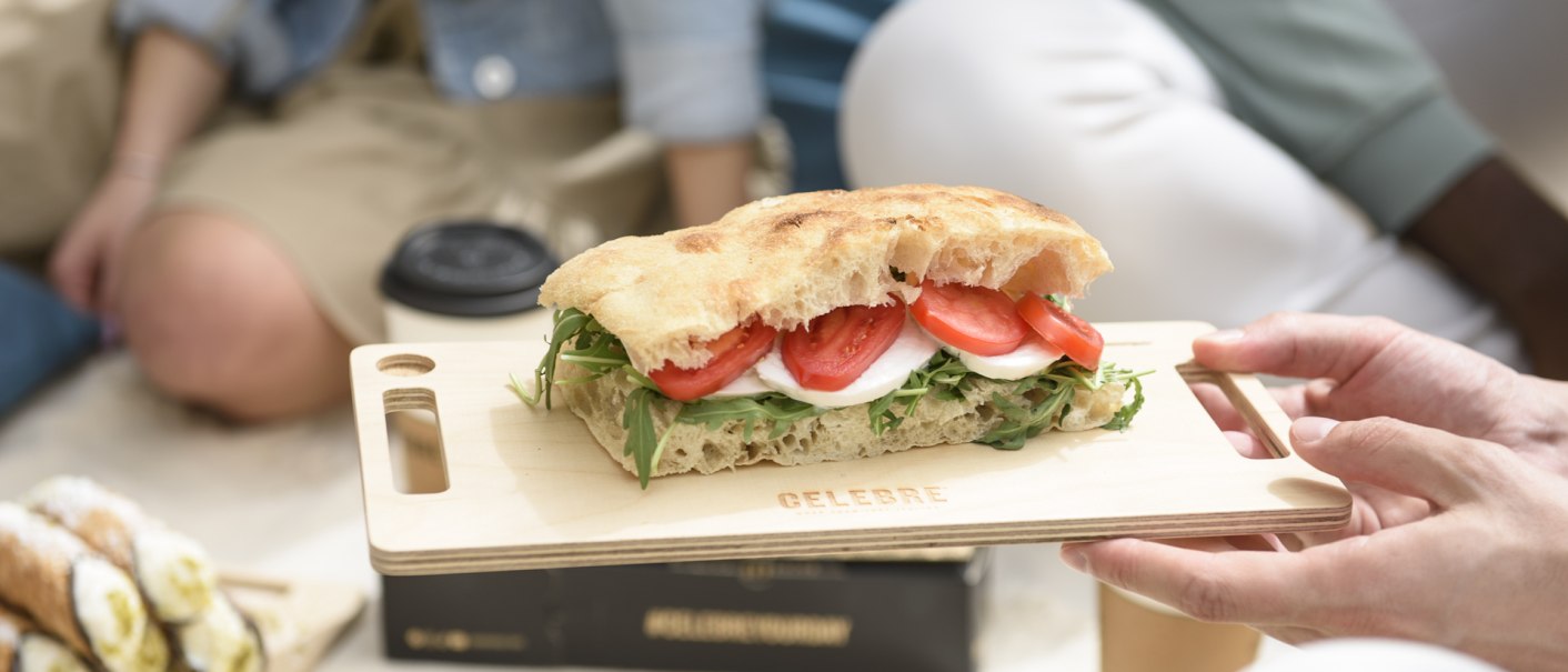 Ein Sandwich mit Tomaten, Mozzarella und Rucola auf einem Holzbrett, gehalten von einer Hand. Im Hintergrund sind Menschen bei einem Picknick zu sehen., © CELEBRE