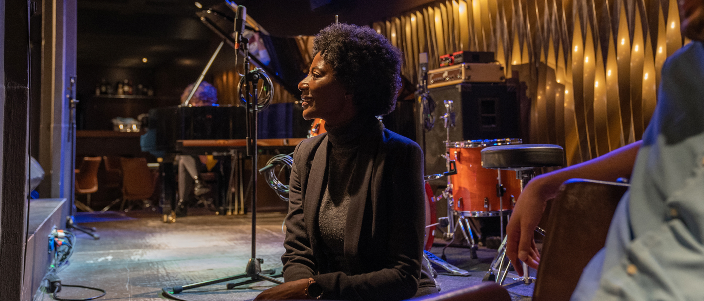 Eine Frau sitzt lächelnd in einem Jazzclub neben einem Schlagzeug. Im Hintergrund ist ein Flügel zu sehen. Die Atmosphäre ist gemütlich und musikalisch., © Stuttgart-Marketing GmbH, Martina Denker