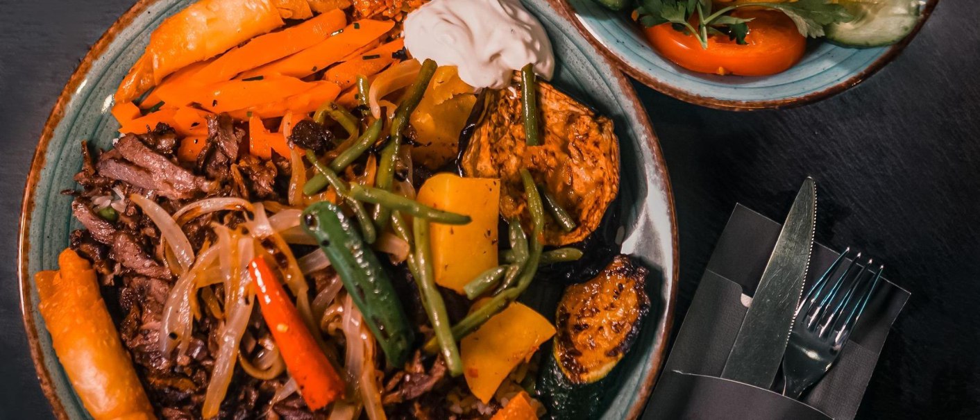 Ein Teller mit gebratenem Fleisch, Gemüse und Joghurt, daneben ein Salat und Besteck auf einem dunklen Tisch., © ALATURKA | Das Stuttgarter Original Ein Teller mit gebratenem Fleisch, Gemüse und Joghurt, daneben ein Salat und Besteck auf einem dunklen Tisch., © ALATURKA | Das Stuttgarter Original