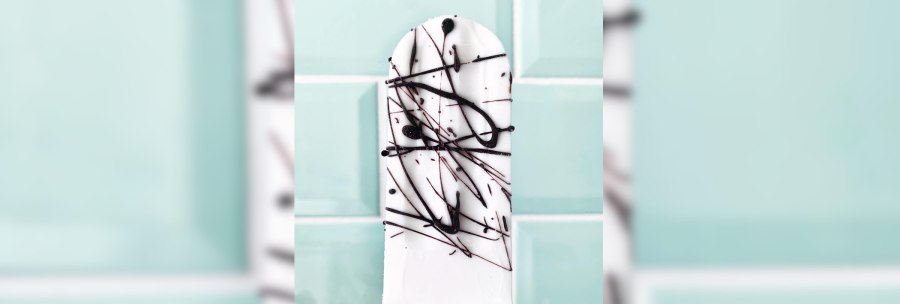 A white popsicle with black chocolate decoration in front of a light blue tiled wall., © Zur Schleckerei A white popsicle with black chocolate decoration in front of a light blue tiled wall., © Zur Schleckerei