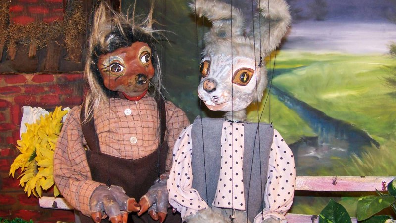 Zwei Marionetten, ein Hase und ein Igel, sitzen auf einer Bank vor einer gemalten Landschaft. Der Hase trägt ein gepunktetes Hemd., © Nellys Puppentheater Zwei Marionetten, ein Hase und ein Igel, sitzen auf einer Bank vor einer gemalten Landschaft. Der Hase trägt ein gepunktetes Hemd., © Nellys Puppentheater