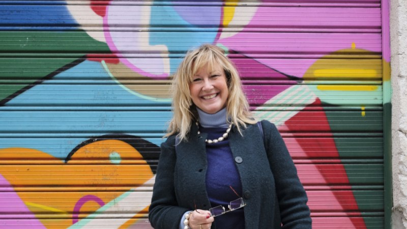 Eine lächelnde Frau steht vor einer bunten Graffiti-Wand. Sie trägt einen dunklen Mantel und hält eine Brille in der Hand., © Foto: privat Eine lächelnde Frau steht vor einer bunten Graffiti-Wand. Sie trägt einen dunklen Mantel und hält eine Brille in der Hand., © Foto: privat