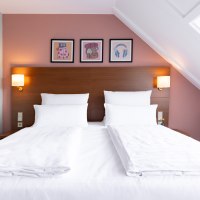 Ein stilvoll eingerichtetes Hotelzimmer mit einem großen Doppelbett, weißen Bettlaken und Kissen. Drei Bilder schmücken die Wand über dem Bett., © Novum Hospitality Ein stilvoll eingerichtetes Hotelzimmer mit einem großen Doppelbett, weißen Bettlaken und Kissen. Drei Bilder schmücken die Wand über dem Bett., © Novum Hospitality