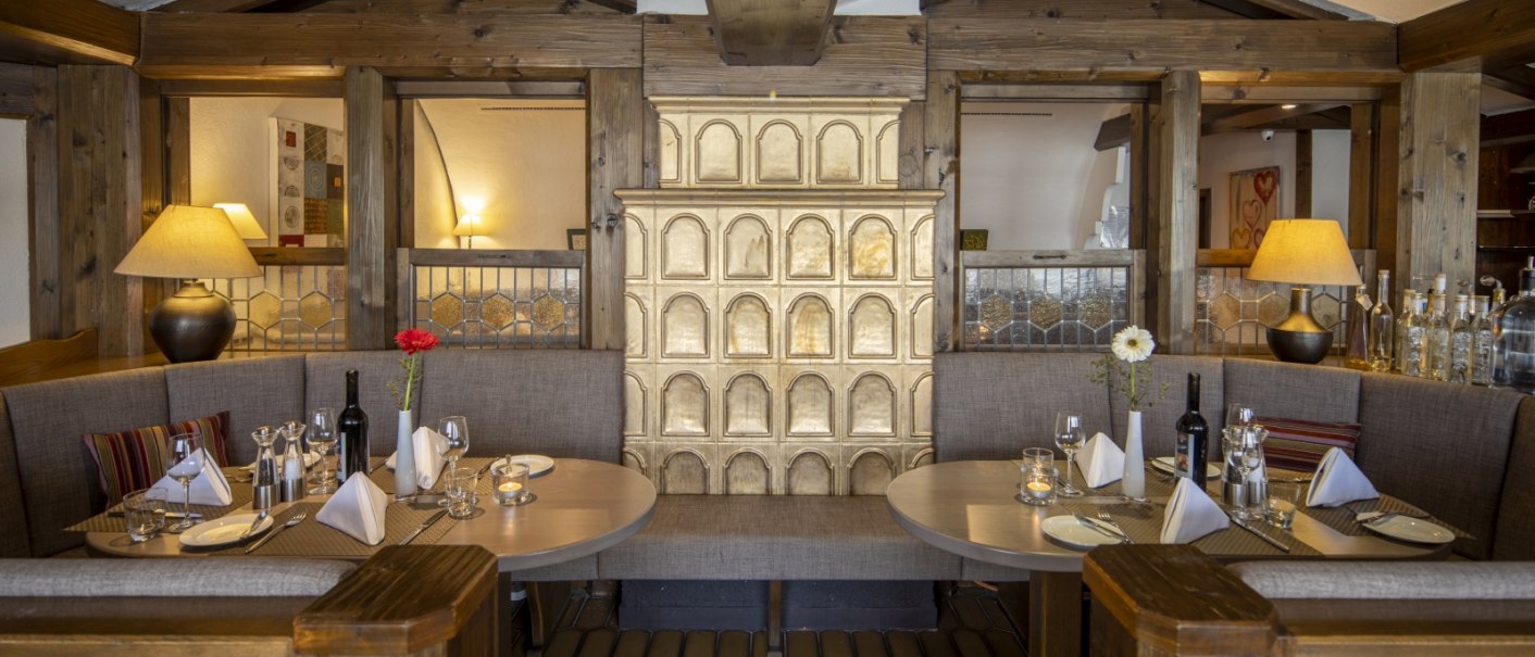 Cozy restaurant corner with wooden tables, covered seating and a decorative tiled stove in the middle., © Hotel Gloria GmbH
