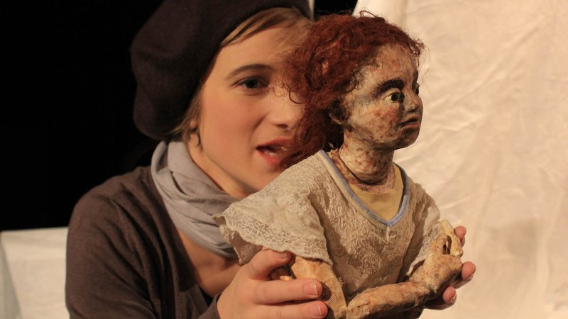 Eine Person mit Mütze hält eine Puppe mit rotem Haar und einem weißen Spitzenkleid vor einem hellen Hintergrund., © FITZ Das Theater animierter Formen Eine Person mit Mütze hält eine Puppe mit rotem Haar und einem weißen Spitzenkleid vor einem hellen Hintergrund., © FITZ Das Theater animierter Formen