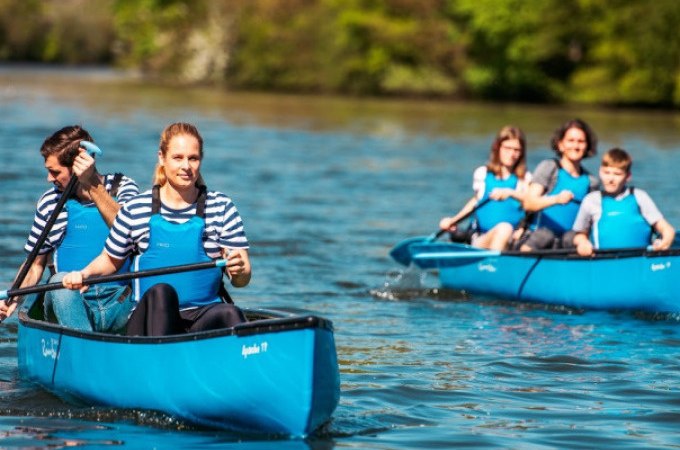 Two blue canoes with people paddling on a river. The people are wearing striped shirts and blue life jackets. There is green vegetation in the background., © Cool-Tours StattReisen Two blue canoes with people paddling on a river. The people are wearing striped shirts and blue life jackets. There is green vegetation in the background., © Cool-Tours StattReisen