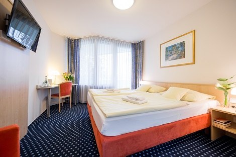 Helles Doppelzimmer mit großem Bett, Schreibtisch, Fernseher an der Wand und Fenster mit Vorhängen. Blumen und Kunstwerk als Dekoration., © Hotel am Schlossberg Herrenberg