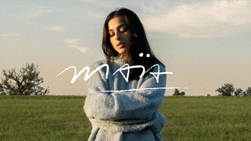 Eine Frau in einem blauen Pullover steht mit geschlossenen Augen auf einer gr&uuml;nen Wiese. Der Schriftzug 'ma&iuml;a' ist prominent im Bild., &copy; links im Bild