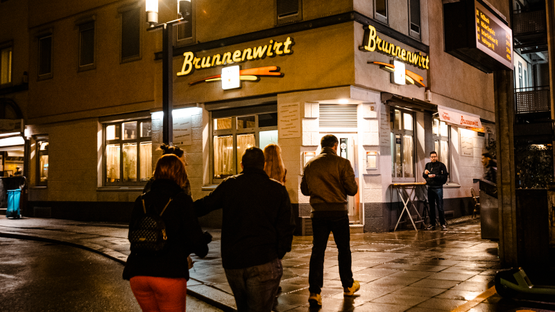 Nachtaufnahme eines Restaurants namens Brunnenwirt. Mehrere Personen stehen davor, die Straße ist nass und ein E-Scooter ist am Rand geparkt., © Stuttgart-Marketing GmbH, Sarah Schmid
