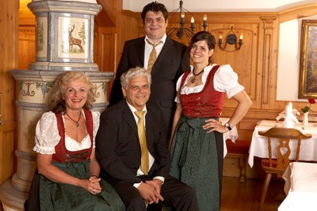 Four people dressed in traditional Bavarian costume are posing in a wood-paneled room with a tiled stove., © Jeanette Bender Four people dressed in traditional Bavarian costume are posing in a wood-paneled room with a tiled stove., © Jeanette Bender
