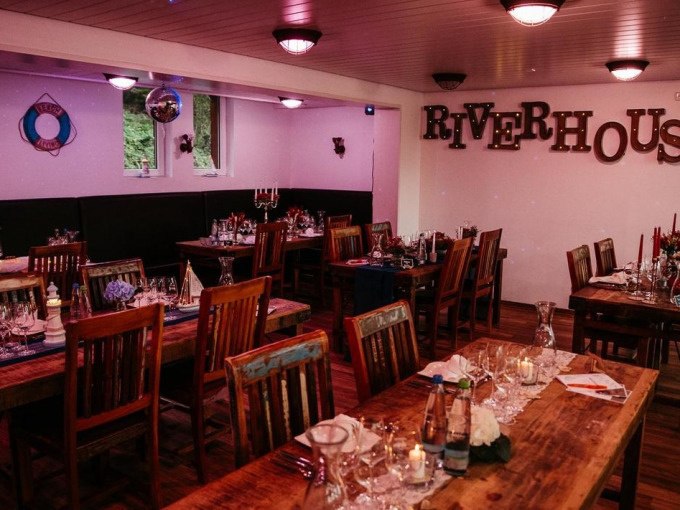 Cozy room with wooden tables, decorated for an event. "RIVERHOUSE" on the wall. Candles and glasses on the tables., © Cool-Tours StattReisen Cozy room with wooden tables, decorated for an event. "RIVERHOUSE" on the wall. Candles and glasses on the tables., © Cool-Tours StattReisen
