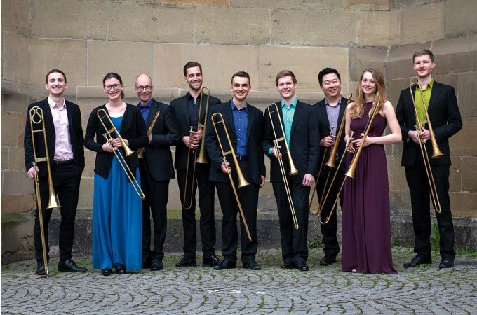 Nine people in festive dress hold trombones and stand in front of a stone wall. They smile and pose for a group photo., &copy; Landesmuseum W&uuml;rttemberg