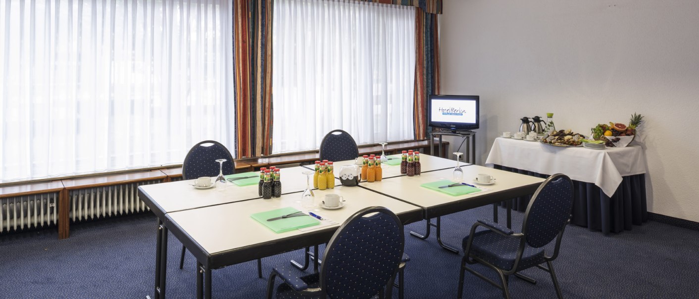 A conference room with a set table, chairs, drinks, and snacks. A screen shows "Hotel Berlin." Windows with curtains in the background., &copy; Hotel Berlin aZIS Hotel BetriebsGmbH