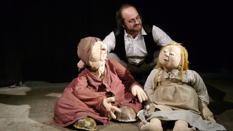 Ein Mann sitzt hinter zwei großen Puppen, die auf dem Boden sitzen. Die Puppen tragen ein braunes und ein graues Kleid. Der Hintergrund ist dunkel., © FITZ Das Theater animierter Formen