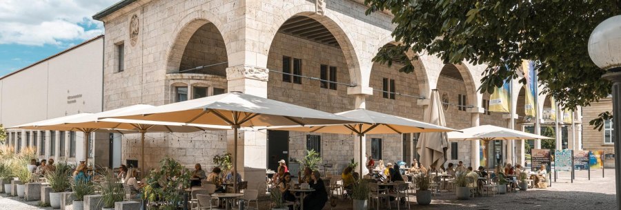 A caf&eacute; with large parasols and guests sitting at tables outside. The building has arches and is made of stone., &copy; Stuttgart Marketing GmbH, Sarah Schmid
