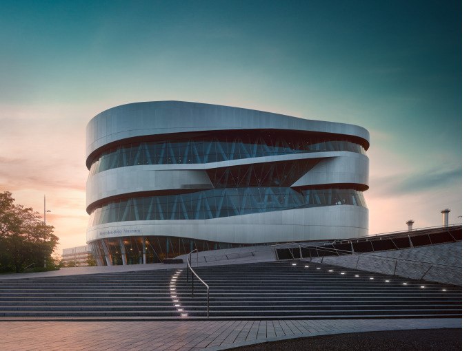 Das Mercedes-Benz Museum mit seiner modernen, geschwungenen Architektur bei Sonnenuntergang, umgeben von Treppen und einem klaren Himmel., &copy; Mercedes-Benz Heritage