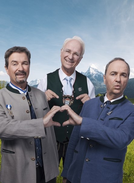 Three men in traditional Bavarian costume stand in front of a mountain backdrop. Two form a heart with their hands, while the third stands smiling in the middle., &copy; Renitenztheater Stuttgart e.V.