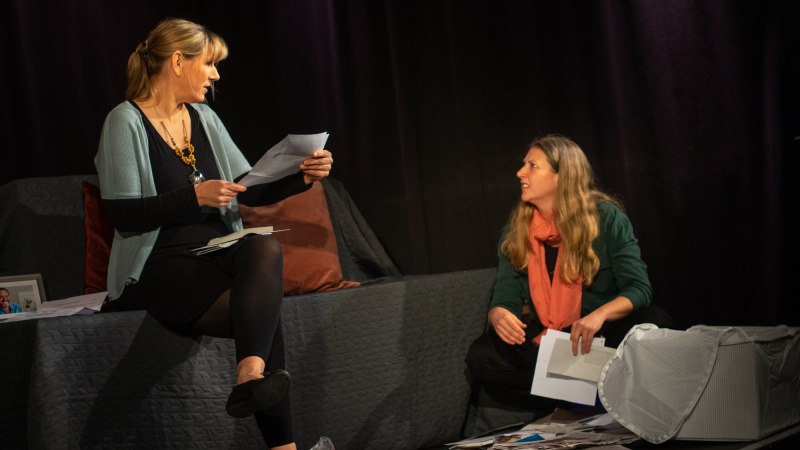Zwei Frauen auf einer B&uuml;hne, eine sitzt auf einem Sofa, die andere auf dem Boden. Sie halten Papiere in der Hand, umgeben von verstreuten Dokumenten., &copy; Improtheater Stuttgart