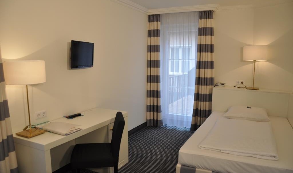Bright single room with bed, desk, chair and wall-mounted TV. Large windows with striped curtains and two floor lamps., &copy; relexa Waldhotel Schatten