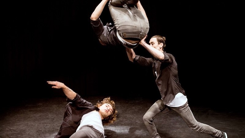 Drei Akrobaten auf einer Bühne: Einer wird in die Luft geworfen, während die anderen beiden ihn stützen. Dynamische Pose vor schwarzem Hintergrund., © Theaterhaus Stuttgart e.V. Drei Akrobaten auf einer Bühne: Einer wird in die Luft geworfen, während die anderen beiden ihn stützen. Dynamische Pose vor schwarzem Hintergrund., © Theaterhaus Stuttgart e.V.