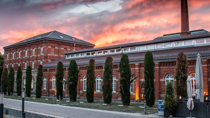 Historisches Backsteingebäude mit Bäumen und Schirmen im Vordergrund, bei dramatischem Sonnenuntergangshimmel., © nestor Hotel Ludwigsburg