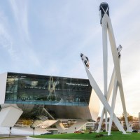 The Porsche Museum with its modern architecture and a sculpture of three cars on high pillars against a blue sky., © Porsche AG, Sabine Braun