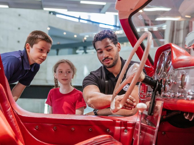 A man explains the cockpit of a red vintage car in a modern museum to two children. The children watch with interest., &copy; Mercedes-Benz Heritage GmbH
