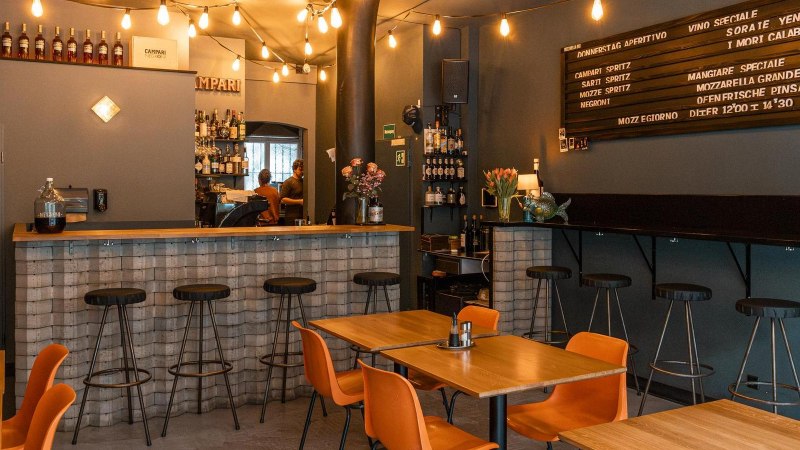 Cozy bar with wooden tables, bar stools and a counter. Fairy lights and a menu board adorn the wall., © SMG, Sarah Schmid Cozy bar with wooden tables, bar stools and a counter. Fairy lights and a menu board adorn the wall., © SMG, Sarah Schmid