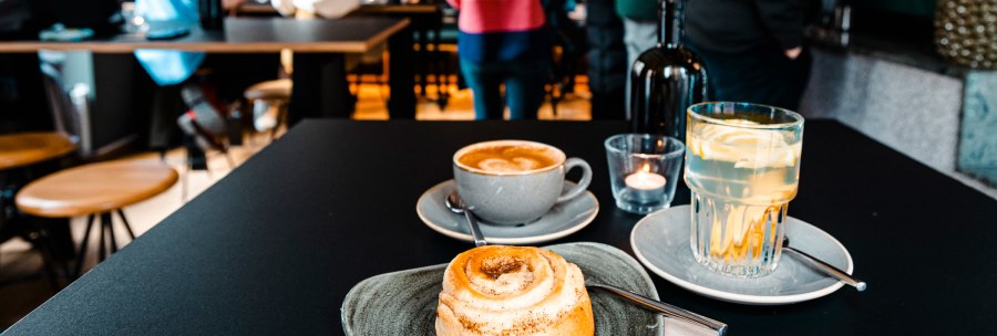 A table in a café with a cinnamon bun, cappuccino and lemon water. Blurred people can be seen in the background., © SMG Stuttgart Marketing GmbH - Sarah Schmid