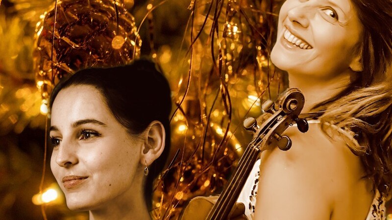 Zwei Frauen mit Geige und Fl&ouml;te l&auml;cheln vor einem festlich geschm&uuml;ckten Weihnachtsbaum. Die Szene ist in warmen, goldenen T&ouml;nen gehalten., &copy; Concerti - Costabel