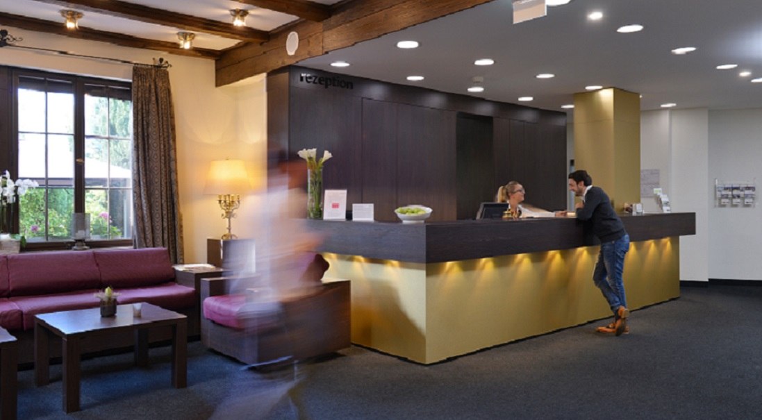 Hotel reception with a man talking to a receptionist. One person walks past quickly. Cozy sitting area with sofa and table., &copy; relexa Waldhotel Schatten