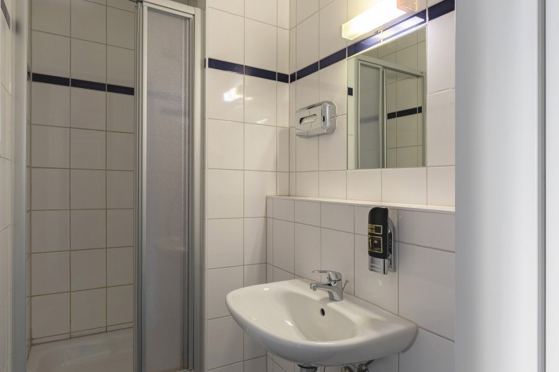 Small, tiled bathroom with shower, washbasin and mirror. A soap dispenser is mounted on the wall., © A&O Hostels Marketing GmbH Small, tiled bathroom with shower, washbasin and mirror. A soap dispenser is mounted on the wall., © A&O Hostels Marketing GmbH