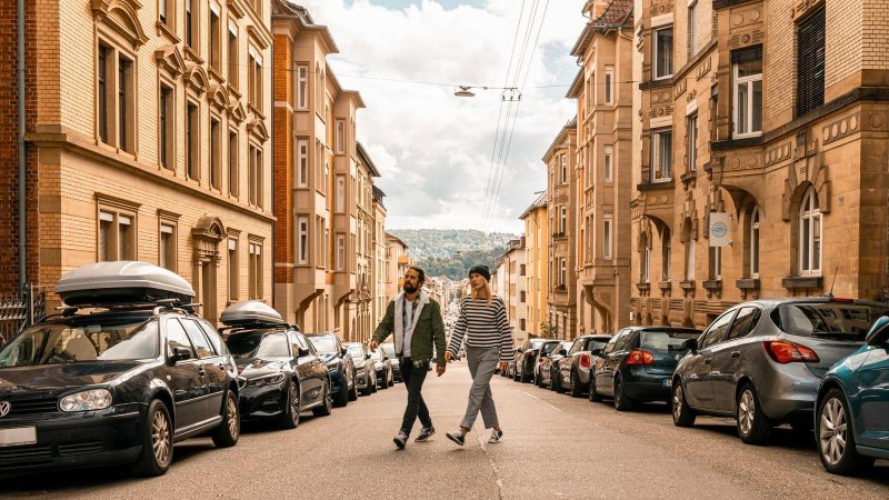 Zwei Personen &uuml;berqueren eine Stra&szlig;e in Stuttgart West. Die Stra&szlig;e ist ges&auml;umt von geparkten Autos und historischen Geb&auml;uden. Der Himmel ist bew&ouml;lkt., &copy; SMG, Sarah Schmid