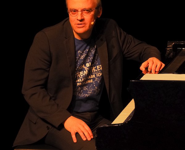 Ein Mann in dunklem Anzug sitzt auf einer Klavierbank, tr&auml;gt ein Mikrofon und spricht. Im Hintergrund ist ein Fl&uuml;gel zu sehen., &copy; Theaterhaus Stuttgart e.V.