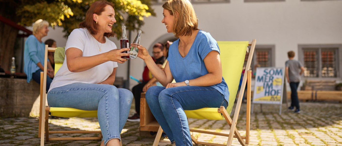 Two women sit on yellow deckchairs and toast with drinks. A sign reading 'Summer in the courtyard' can be seen in the background., © FTGRF.de