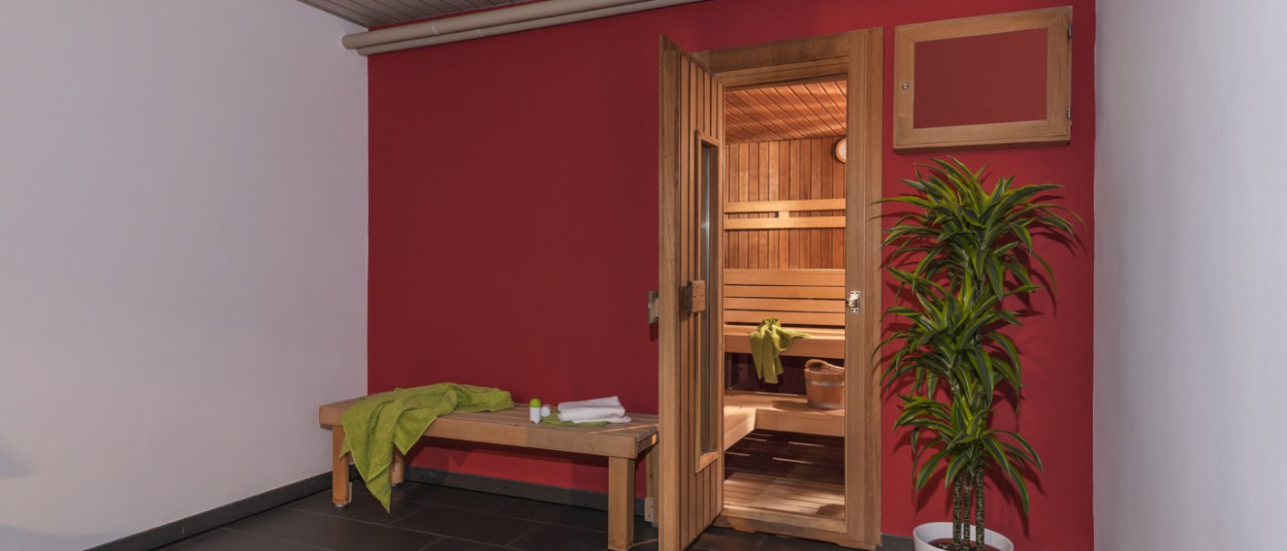 Sauna area with a red wall, an open sauna door, a wooden bench with green towels, and a plant in a white pot., &copy; Hotel Berlin aZIS Hotel BetriebsGmbH