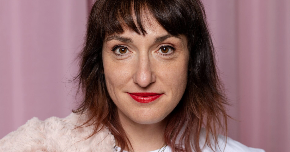 Nahaufnahme einer Frau mit braunen Haaren und rotem Lippenstift vor einem rosa Hintergrund. Sie trägt ein weißes Oberteil und einen flauschigen Schal., © Julia Ruhland Nahaufnahme einer Frau mit braunen Haaren und rotem Lippenstift vor einem rosa Hintergrund. Sie trägt ein weißes Oberteil und einen flauschigen Schal., © Julia Ruhland