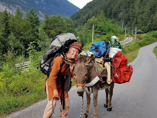Eine Person mit großem Rucksack steht lächelnd neben einem beladenen Esel auf einer Bergstraße, umgeben von grünen Wäldern und Bergen., © Theaterhaus Stuttgart e.V. Eine Person mit großem Rucksack steht lächelnd neben einem beladenen Esel auf einer Bergstraße, umgeben von grünen Wäldern und Bergen., © Theaterhaus Stuttgart e.V.
