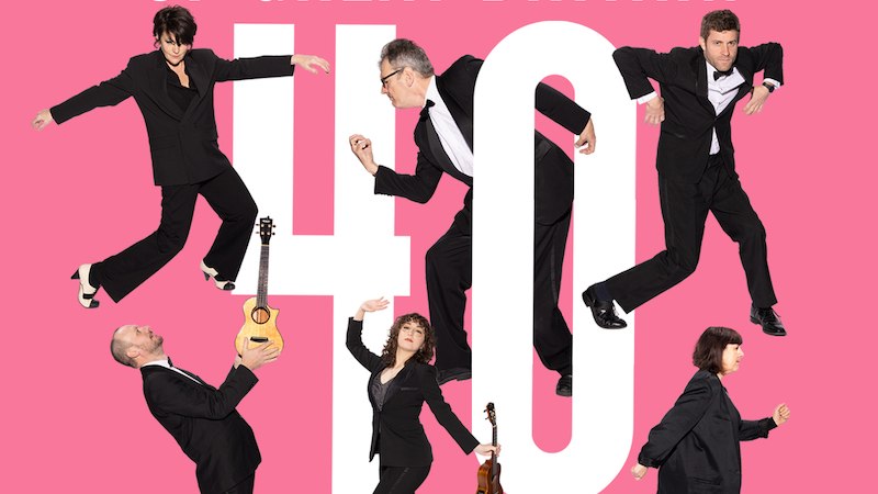 Six people in suits pose on a pink background with the inscription 'Ukulele Orchestra of Great Britain 40 Years'., &copy; Theaterhaus Stuttgart e.V.