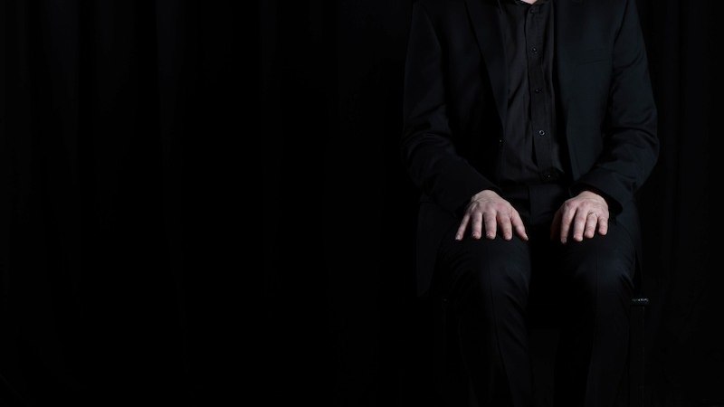 Person in schwarzem Anzug sitzt auf einem Stuhl vor schwarzem Hintergrund, schaut ernst in die Kamera., © Renitenztheater Stuttgart e.V. Person in schwarzem Anzug sitzt auf einem Stuhl vor schwarzem Hintergrund, schaut ernst in die Kamera., © Renitenztheater Stuttgart e.V.